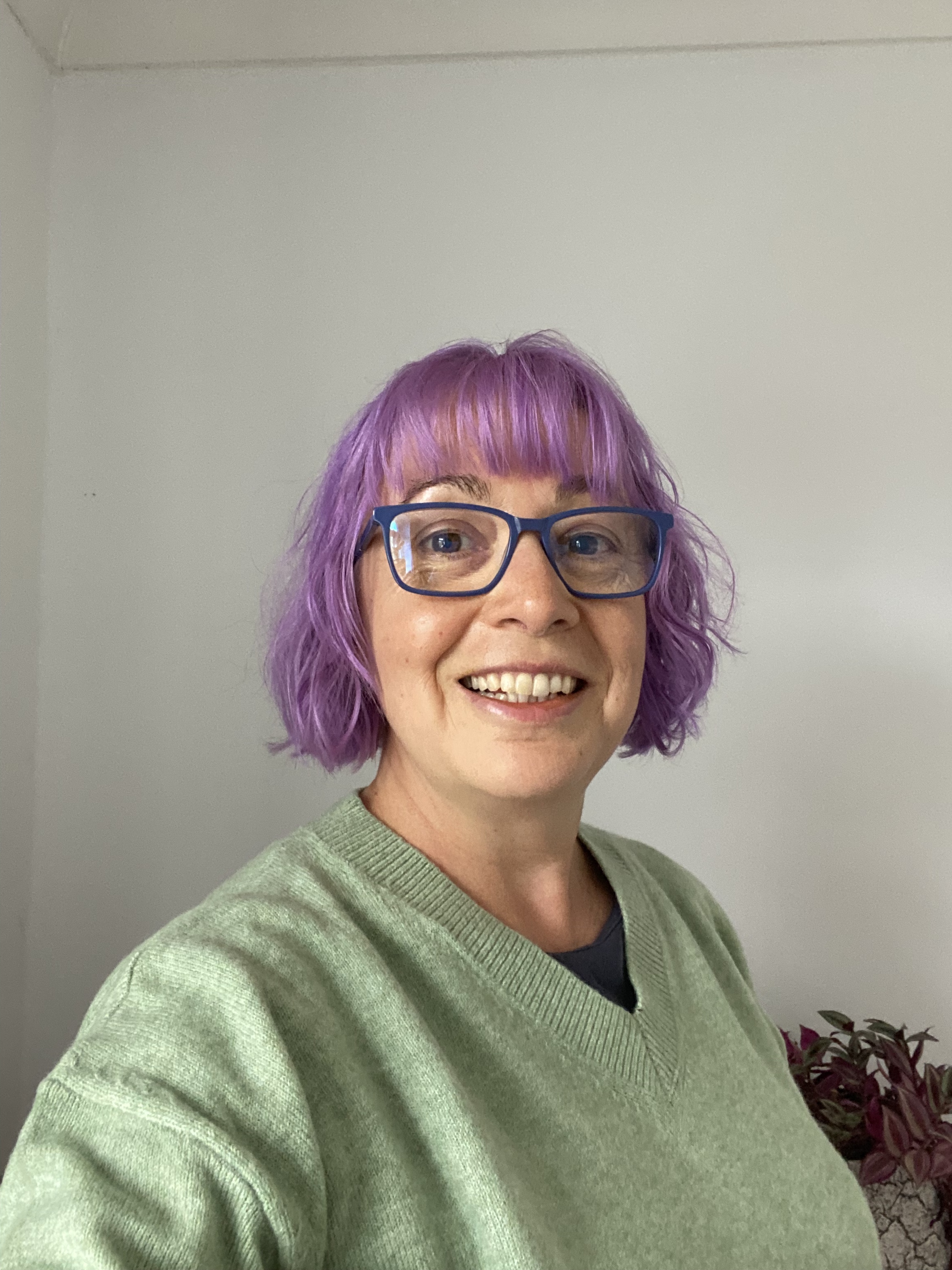 Woman with purple hair, blue glasses and wearing a light green jumper is looking at the camera and smiling